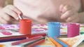 Table filled with acrylic paint and brushes, children drawing in background HD Footage