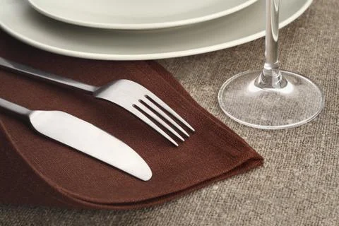Table setting. Cutlery set with fork, knife, plate and glass on brown linen napk Stock Photos