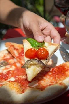 At the table, someone picks up a slice of pizza with tomatoes and basil. Round Stock Photos