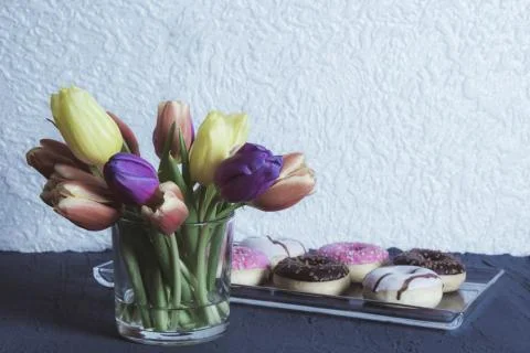 On the table there is a vase with tulips in the back are delicious donuts Stock Photos