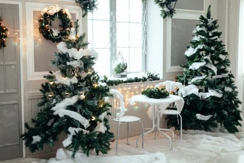Table with two chairs between Christmas trees covered with snow and garlands  库存照片