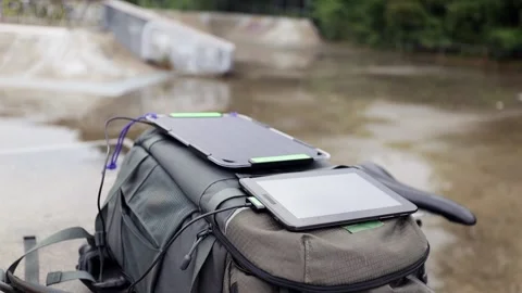 Tablet is charged from a portable solar panel battery attached to backpack. 스톡 동영상 157333253