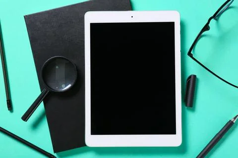Tablet computer with notebook, eyeglasses and magnifying glass on turquoise b Stock Photos