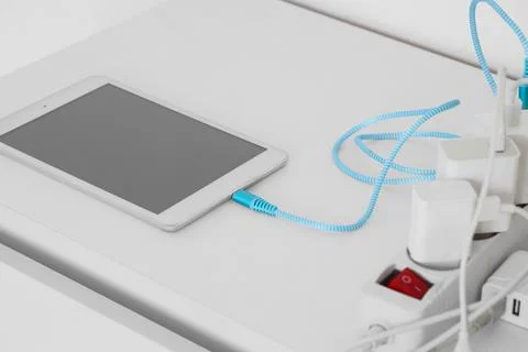 Tablet connected to power strip with blue charging cable on white desk Stock Photos