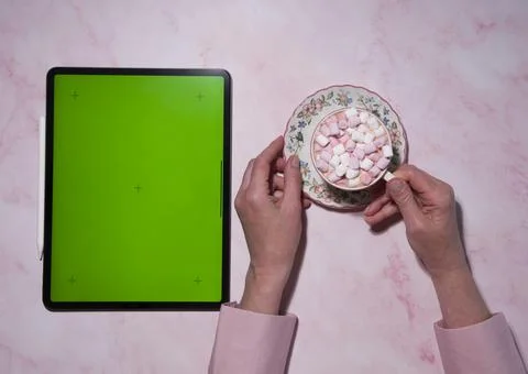 A tablet with a green screen is on the table, next to a woman is holding a cup Stock Photos