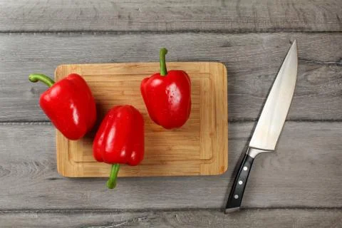 Tabletop view - three whole red bell peppers on chopping board, knife next to Stock Photos