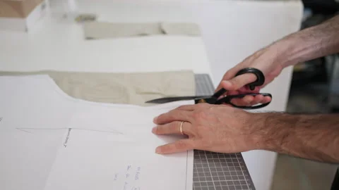 Tailor cutting fabric with scissors following paper pattern on worktable Stock Footage 327653794