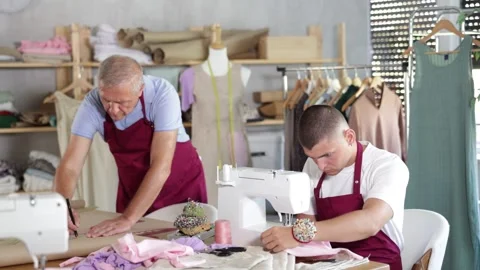 Tailor sits at a table and sews on a machine next to an experienced worker in a Stock Footage 332637994