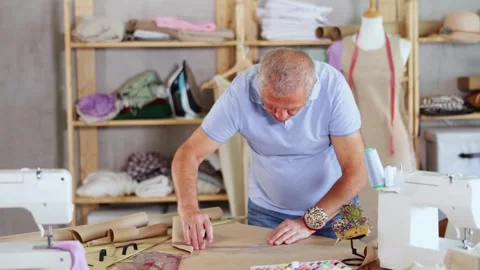 Tailor stands near the table and draws patterns of clothes with a pencil on Stock Footage 325772225