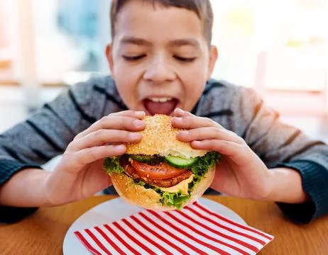 Taking a huge bite. a young boy eating a delicious burger while sitting at his Stock Photos