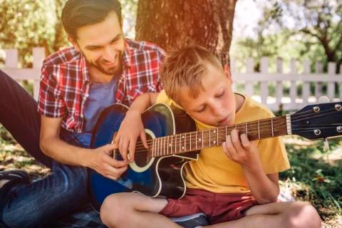 Talented boy is looking on strings and playing guitar. His father is sitting Stock Photos