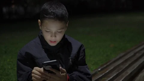 Talkitive kid playing mobile game on the bench outdoors at night, close up Vidéo 98878339