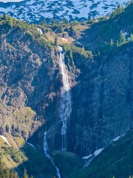 Tall Alpine Waterfall Cascading Down Rocky Mountain Cliff Amid Snow Patches.. Stock Photos