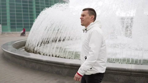 A tall man in a white jacket walks near the fountain in rainy weather Stock Footage 218979166