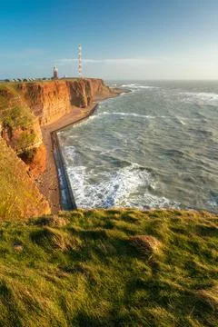 Tall red dramatic cliffs of Heligoland island with rough sea and a lighthouse Stock Photos