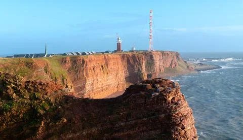 Tall red dramatic cliffs of Heligoland island with rough sea and a lighthouse Stock Photos