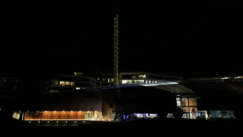 Tall spiral lit tower rises into black sky above wood clad modern buildings Stock Footage 326358702