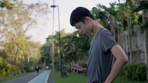 A tall, thin, black-haired young Asian man playing smartphone at the park. Stock Footage 162081122