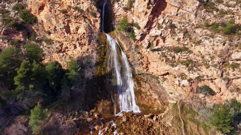 Tall waterfall cascading down rugged orange rocky cliffs surrounded by green 库存影片 334785055
