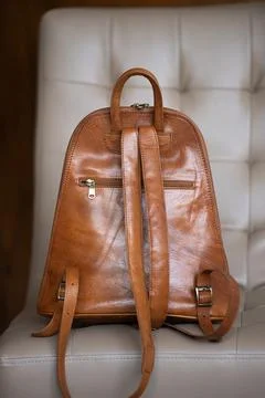 A tan leather backpack with subtle detailing rests on a light cream tufted chair Stock Photos