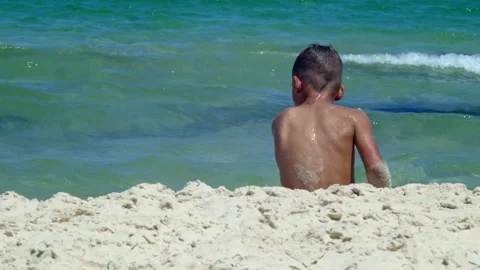 A tanned boy is sitting on the white sand by the sea with his back to the camera Stock Footage 157998627