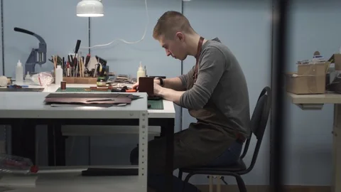 A tanner in an apron sits at a table in the workshop and punches holes in Stock Footage 104925305