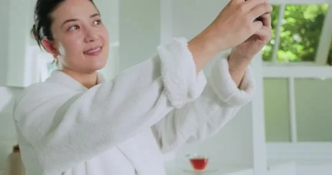 Tapping screen, woman in white plush robe raising black phone at bathroom Stock Footage 334925209
