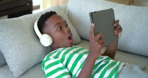 Tapping tablet once African American boy on gray couch wearing headphones Stock Footage 340881558