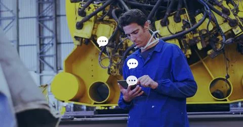 Tapping tech in blue coverall checking phone at factory, with yellow machine and Stock Photos