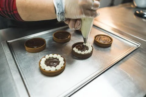 Tarts placed on a baking tray being decorated with white cream by a person using Stock Photos