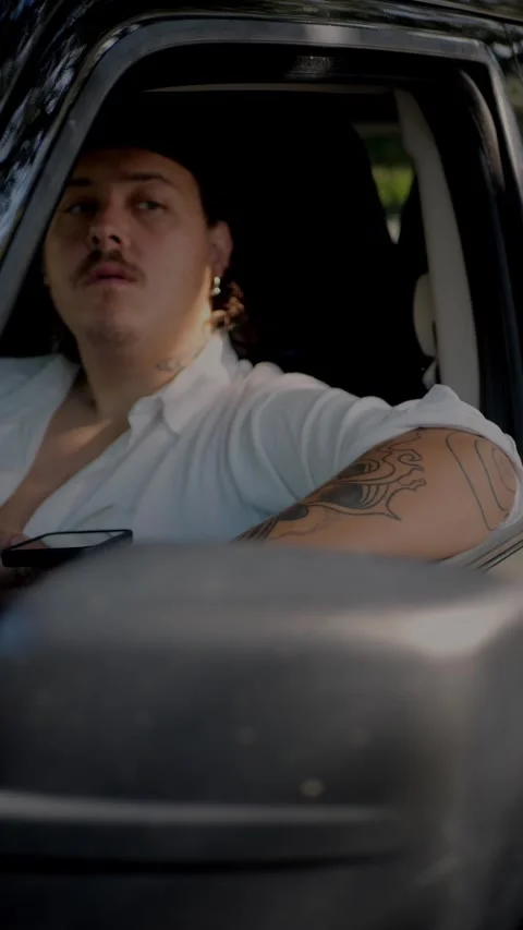 Tattooed man sitting in car with window open, holding smartphone... Stock Footage 324632472
