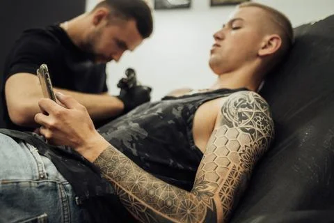 Tattooed Man Using Smartphone During Tattoo Session, Male Artist Draws on the Stock Photos
