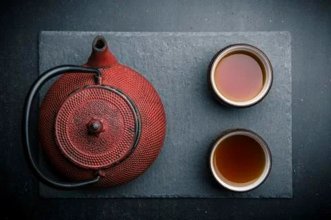 Tea composition with ceramic tea cups and red iron teapot on dark stone backg Stock Photos