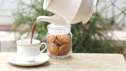 Tea pot pouring Tea in tea cup and saucer with cookies in background Video stock 197746192