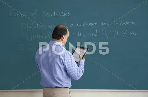 Teacher With Digital Tablet Writing On Blackboard
