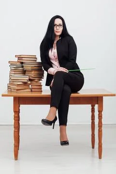 A teacher with a school pointer sits on a table with many books Stock Photos