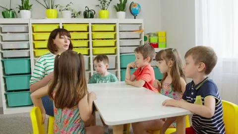 The teacher sits down and explains the task to the children. Vídeos de archivo 251752099