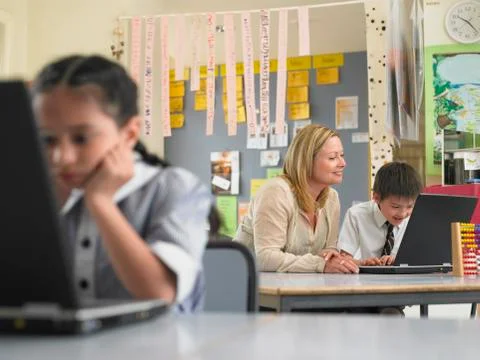 Teacher Sitting With Boy Using Laptop In Class Stock Photos