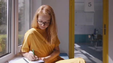 Teacher sitting at school window, writing in notebook and smiling at the camera 스톡 동영상 162735547