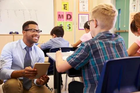 Teacher using tablet computer kneeling beside pupil's desk 스톡 사진