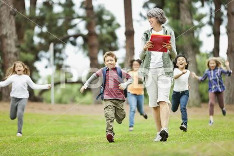 Teacher Walking At Park While Students Run In Background Stock Photo 