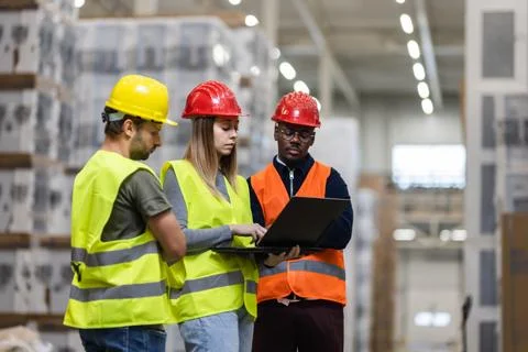 Team discussing logistics in warehouse using laptop and protective gear Foto stock