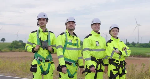 Team of engineers in bright uniform slowly standing under wind turbines, 스톡 동영상 330761870