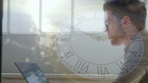 Tech man working on laptop beside window, sunlight rising causing clock hands Stock Footage 335235833
