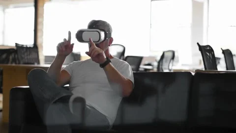 Tech worker seated on sofa with VR HMD testing VR interface on red particles Stock Footage 331872633
