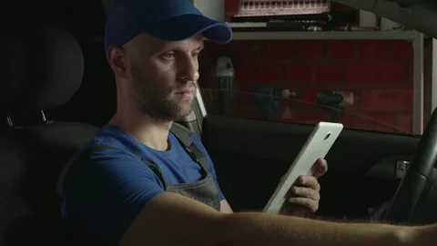 A technician is inside a car in a garage, looking at a tablet in his hands. He Stock-Footage 280378538