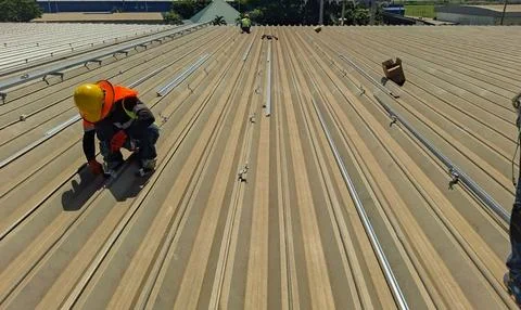 Technicians installing solar panel mounting rails on the metal roof of an ind Stock Photos