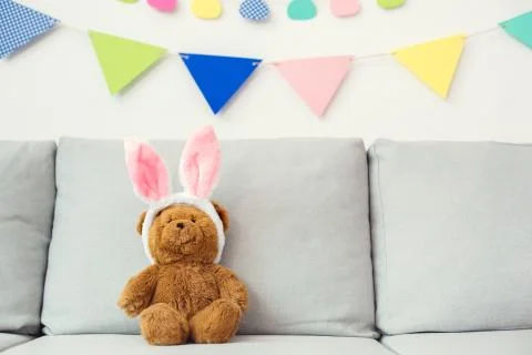 Teddy bear sitting on the sofa in bunny ears bright background Stock Photos
