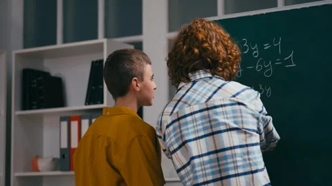 Teen boy helping classmate with a math problem near chalkboard, algebra lesson Stock Footage 251805496