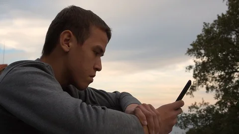 Teen Boy Sits Alone, Using a Smartphone, Looking Sad Or Worried Stock Footage 97629257